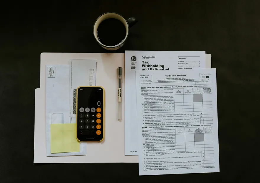 A workspace with tax forms, a calculator, a pen, an envelope, sticky notes, and a cup of coffee on a dark desk.