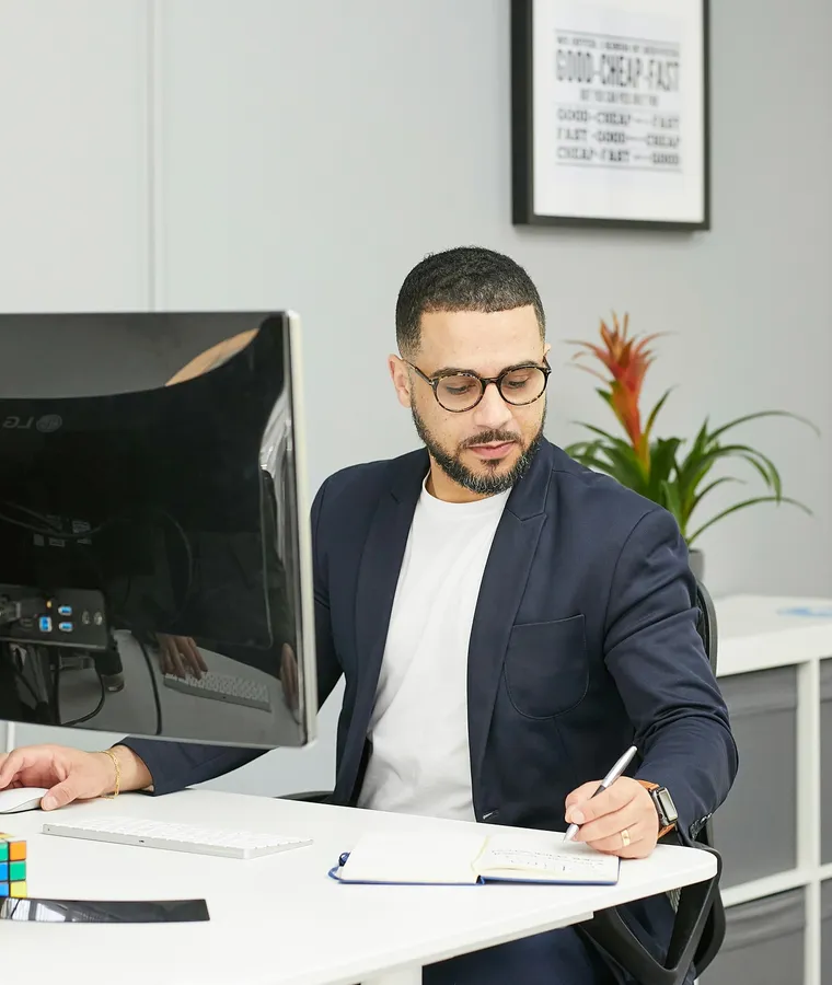 Modern bir ofiste, bilgisayar monitörünün yanında not defterine yazan gözlüklü ve ceketli bir adam.