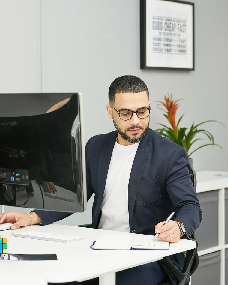 Modern bir ofiste, bilgisayar monitörünün yanında not defterine yazan gözlüklü ve ceketli bir adam.