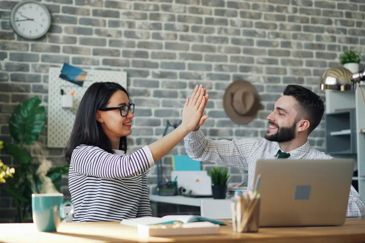 Two colleagues smiling and giving a high-five at a desk in a modern office with a laptop, coffee mug, and plants.