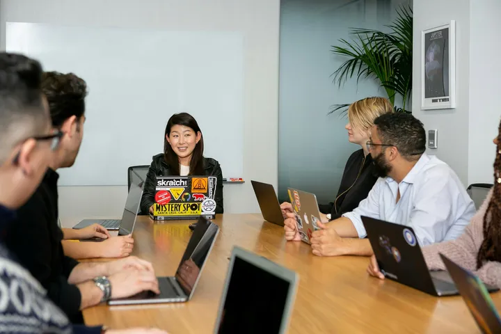 A group of colleagues sitting around a conference table with laptops, listening to a woman leading the meeting in a modern office setting.