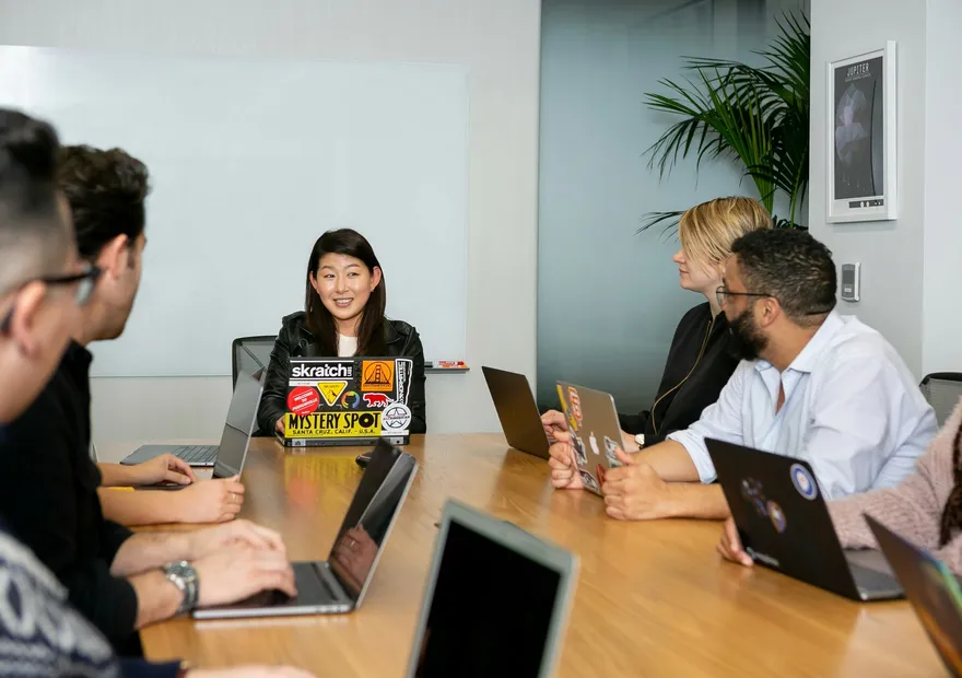 A group of colleagues sitting around a conference table with laptops, listening to a woman leading the meeting in a modern office setting.
