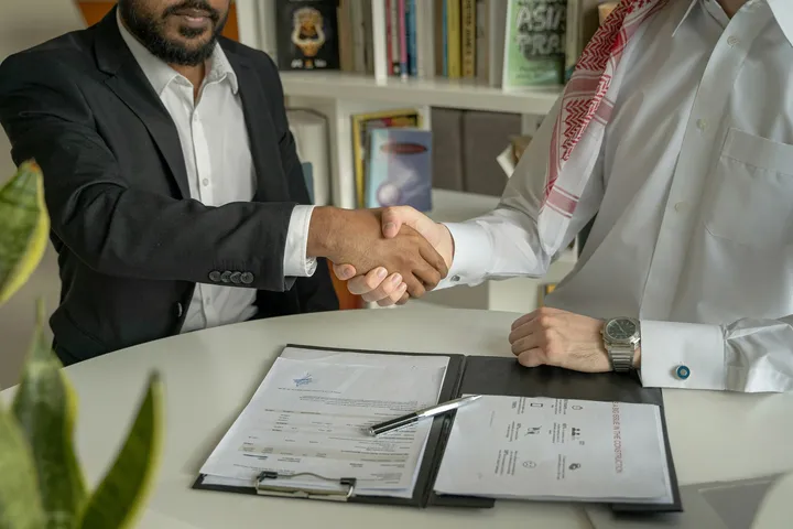 Two businessmen shaking hands across a desk with documents and a pen, symbolizing a successful business agreement.