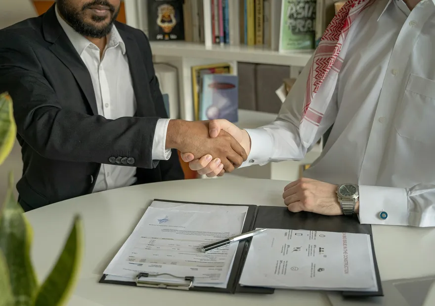 Two businessmen shaking hands across a desk with documents and a pen, symbolizing a successful business agreement.